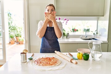 Poster - Beautiful blonde woman wearing apron cooking pizza shocked covering mouth with hands for mistake. secret concept.