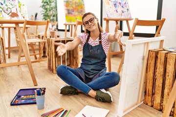 Canvas Print - Young brunette woman at art studio sitting on the floor looking at the camera smiling with open arms for hug. cheerful expression embracing happiness.