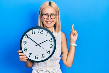 Poster - Beautiful blonde woman holding big clock smiling with an idea or question pointing finger with happy face, number one