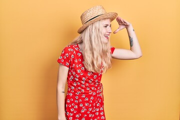 Canvas Print - Beautiful caucasian woman with blond hair wearing summer hat very happy and smiling looking far away with hand over head. searching concept.