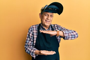 Poster - Handsome senior man with grey hair wearing welding protection mask gesturing with hands showing big and large size sign, measure symbol. smiling looking at the camera. measuring concept.