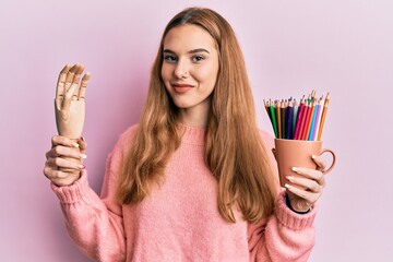 Sticker - Young blonde woman holding small wooden manikin hand and pencils smiling with a happy and cool smile on face. showing teeth.