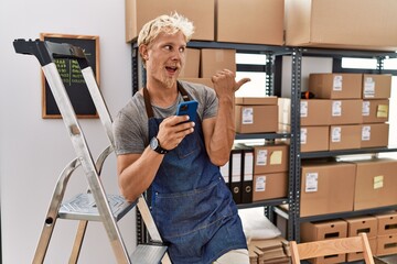 Poster - Young blond man using smartphone working at storehouse smiling with happy face looking and pointing to the side with thumb up.