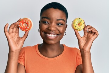 Sticker - Young african american woman holding tasty colorful doughnuts on eyes winking looking at the camera with sexy expression, cheerful and happy face.