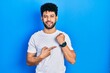 © Krakenimages.com - Young arab man with beard wearing casual white t shirt in hurry pointing to watch time, impatience, looking at the camera with relaxed expression