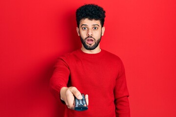 Poster - Young arab man with beard holding television remote control afraid and shocked with surprise and amazed expression, fear and excited face.