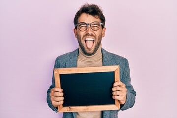 Poster - Handsome man with beard holding blackboard sticking tongue out happy with funny expression.