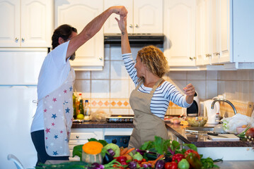 Joyful couple in apron holding hands and dancing together in domestic kitchen at home. Man and woman having fun dancing wearing apron in kitchen. Dancing caucasian couple with raw vegetable on kitchen