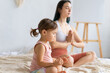 © Вадим Каштанов - Young mom practices yoga on couch with her little daughter. Lotus position meditation. Padmasana exercise.