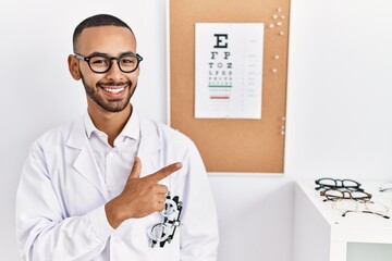 Sticker - African american optician man standing by eyesight test cheerful with a smile on face pointing with hand and finger up to the side with happy and natural expression