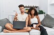 © Krakenimages.com - Young interracial couple using laptop at home sitting on the sofa pointing up looking sad and upset, indicating direction with fingers, unhappy and depressed.