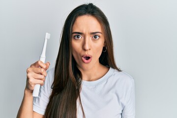 Sticker - Young hispanic girl holding electric toothbrush scared and amazed with open mouth for surprise, disbelief face