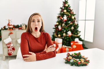 Canvas Print - Caucasian young blonde woman sitting on the table by christmas tree afraid and shocked with surprise expression, fear and excited face.