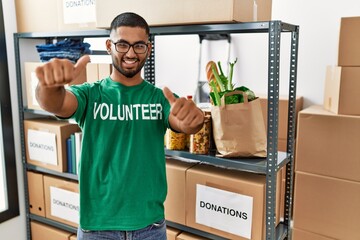 Poster - Young indian man volunteer holding donations box approving doing positive gesture with hand, thumbs up smiling and happy for success. winner gesture.