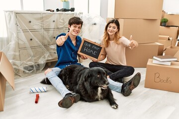 Young caucasian couple with dog holding our first home blackboard at new house approving doing positive gesture with hand, thumbs up smiling and happy for success. winner gesture.