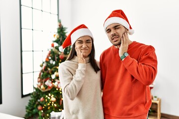 Young hispanic couple standing by christmas tree touching mouth with hand with painful expression because of toothache or dental illness on teeth. dentist concept.