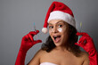 Sexy pretty woman in Santa hat, wears under eyes patches, rejoices looking at camera, holds syringes with beauty injection against gray background. Copy space for Christmas ad. Anti-aging cosmetology