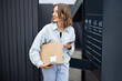 © rh2010 - Young caucasian woman with parcel near automatic post terminal. Smiling girl holding smartphone and looking away on city street. Concept of smart delivery. Idea of modern shipping and logistics
