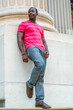 © Alexander Image - Wearing a red, pink stripe Henley V Neck T shirt,  gray pants, leather sneakers, a young black guy is standing against the base of a column, bending over a leg, waiting for you..