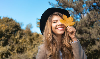 stylish smiling young woman covering her eyes with yellow autumn leaf in park