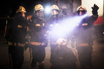 Wall Mural - Group of professional firefighters posing. Firemen wearing uniforms, protective helmets and oxygen masks. Smoke and firetrucks in the background.