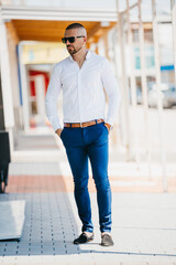 Wall Mural - Cool handsome Caucasian man wearing an elegant white shirt and sunglasses posing outdoor