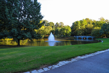 a gorgeous shot of the silky green lake water with a fountain in the middle of the lake with a stone bridge surrounded by lush green and autumn colored trees at Lenox Park in Brookhaven Georgia USA