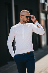 Wall Mural - Cool handsome Caucasian man wearing an elegant white shirt and sunglasses posing outdoor