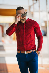 Wall Mural - Cool handsome Caucasian man wearing an elegant red shirt and sunglasses posing outdoor