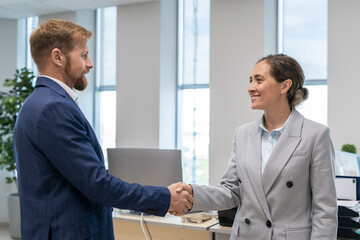 Wall Mural - Two happy business partners in elegant suits shaking hands against workplace in office