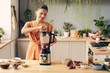© pressmaster - Young woman mixing ingredients of smoothie in electric blender while preparing healthy drink