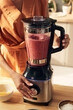 © pressmaster - Young woman pressing button of electric blender while mixing and shaking smoothie ingredients by kitchen table