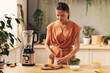 © pressmaster - Young woman preparing ingredients for homemade smoothie while standing by kitchen table