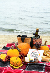 Pumpkins, tasty food, glass of wine and card with text HAPPY THANKSGIVING on beach near water