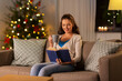 © Syda Productions - winter holidays, leisure and people concept - young woman reading book at home in evening over christmas tree lights on background