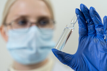 Wall Mural - A woman doctor in glasses and a medical mask looks at a glass ampoule with a transparent liquid in her hands. Medicine development concept. Close-up, blurred background.