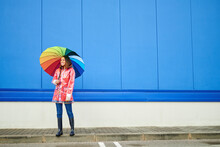 Woman With Multi Colored Umbrella Standing On Footpath By Blue Wall