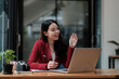 © Natee Meepian - Image of happy asian woman taking note and waving hand at laptop, while speaking or chatting on video call in office