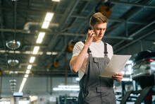Low-angle View Of Focused Handsome Young Mechanic Male Wearing Uniform Reading Clipboard And Talking On Mobile Phone, Standing In Auto Repair Shop Garage, With Vehicle Background, Looking At Camera