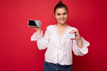 Young beautiful european stylish brunette woman wearing white blouse and fashionable hat isolated over red background with positive sincere emotions. Simple and natural looking at the camera. Holding