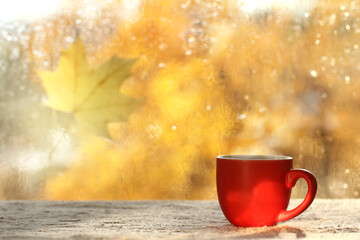 red cup on the table against the background of a wet window with a blurred silhouette of a maple leaf. sunny autumn morning with coffee