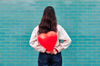 © VISTA by Westend61 - Young woman holding red heart shape balloon behind her back in front of brick wall