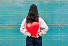 Young Woman Holding Red Heart Shape Balloon Behind Her Back In Front Of Brick Wall