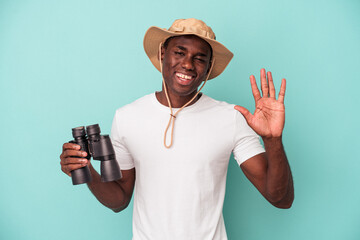 Wall Mural - Young African American man holding binoculars isolated on blue background smiling cheerful showing number five with fingers.