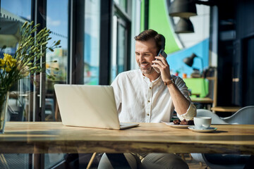 Happy man working using laptop and talking on the phone in a cafe