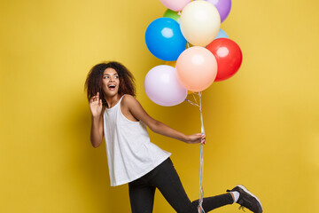 Wall Mural - Celebration Concept - Close up Portrait happy young beautiful african woman with white t-shirt running with colorful party balloon. Yellow Pastel studio Background