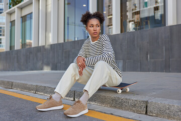 People youth generation and hobby concept. Thoughtful millennial girl with two hair buns rests at longboard dressed in streetwear enjoys recreation chill poses at urban setting. Stylish skateboarder