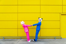 Man And Woman Wearing Vibrant Suits And Bear Masks Holding Hands Against Yellow Wall