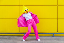 Woman Wearing Vibrant Pink Suit And Bird Mask Standing In Front Of Yellow Wall With Spread Jacket