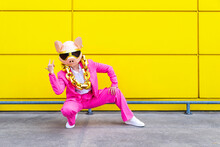 Woman Wearing Vibrant Pink Suit, Pig Mask And Large Golden Chain Crouching In Front Of Yellow Wall